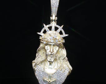 Può includere: Un ciondolo color oro raffigurante il busto dettagliato di una figura con capelli lunghi, che indossa una corona di spine e un'aureola. Il pezzo è incastonato con numerose pietre chiare, inclusa una croce centrale e una contromaglia decorata.