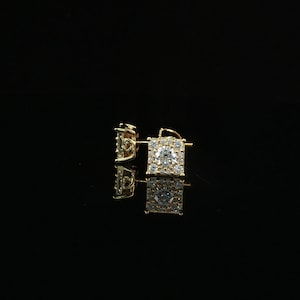 May include: A pair of square-shaped gold stud earrings featuring a central round brilliant-cut clear stone surrounded by a halo of smaller pavé-set clear stones. The earrings have a post backing and are displayed against a solid black background.