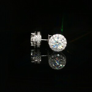 May include: A pair of silver-toned stud earrings featuring a large, round, faceted clear stone at the center, surrounded by a halo of smaller, sparkling clear stones. The side of the setting is also encrusted with a row of tiny clear stones.