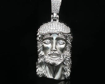 Può includere: Un ciondolo color argento che raffigura il volto dettagliato e tridimensionale di Gesù con capelli lunghi e barba. La parte superiore della testa è decorata con una superficie pavé di piccole pietre chiare e anche la contromaglia è incastonata.
