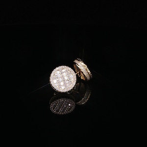 May include: A pair of circular stud earrings featuring a gold-tone metal setting encrusted with numerous small, clear, sparkling stones arranged in a grid pattern across the face and along the thick, rounded edges of each earring piece.