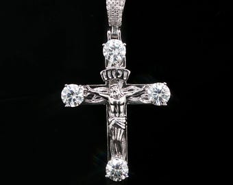 Può includere: Un ciondolo a crocifisso in metallo color argento con la figura centrale di Gesù. La croce è decorata con quattro grandi pietre rotonde e trasparenti sfaccettate alle estremità di ogni braccio e una contromaglia incastonata di pietre.