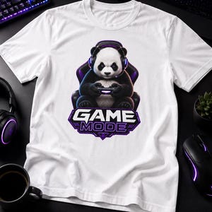 May include: A white short-sleeve t-shirt featuring a graphic of a panda wearing gaming headphones and sitting in a gaming chair while holding a controller. Below the panda, the text "GAME MODE" is printed in a bold, stylized font.