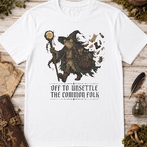 Puede incluir: Camiseta blanca con una ilustración de fantasía de una rana mago, con un bastón. El texto "OFF TO UNSETTLE THE COMMON FOLK" está impreso debajo, creando un diseño único.