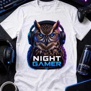 May include: A white t-shirt featuring a graphic of a brown owl wearing black headphones and glasses. Below the owl, the text reads NIGHT GAMER in white and blue capital letters. The owl has bright orange eyes and a detailed feather pattern.