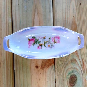 May include: Oval-shaped, vintage porcelain dish with handles. The dish features a white base with a pink and white floral design. The rim has a light purple iridescent finish. The dish is set on a wooden surface.