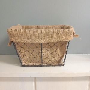 May include: A rectangular wire basket with a burlap liner. The basket has a dark metal frame and a chicken wire design. The burlap liner is folded over the top edge of the basket.