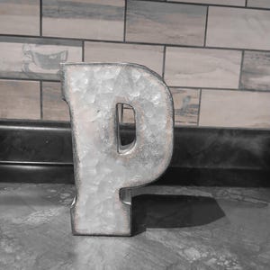 Puede incluir: Una letra "P" metálica con un acabado galvanizado envejecido. La letra es independiente y tiene una superficie texturizada. El fondo presenta una pared de azulejos y una encimera oscura.