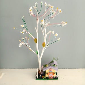 May include: A decorative white tree with small white flowers, green, pink, and blue wire accents, and a miniature house. The house has a purple roof, a yellow door with the word "Easter", and is surrounded by greenery.