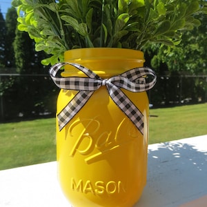 May include: A yellow mason jar with a black and white checkered ribbon tied around the top. The jar is filled with green leafy plants.