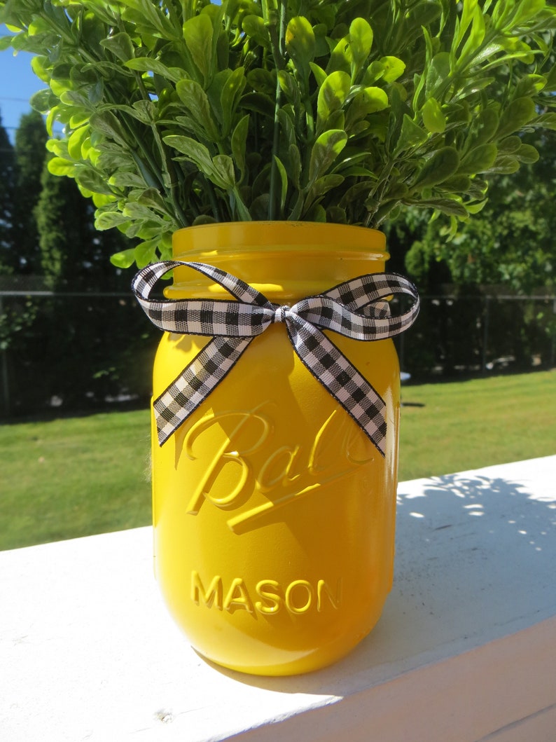 May include: A yellow mason jar with a black and white checkered ribbon tied around the top. The jar is filled with green leafy plants.