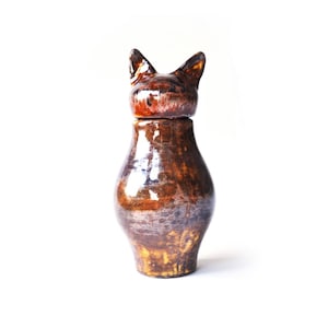 May include: A handcrafted ceramic cat-shaped jar with a lid. The jar is a rich brown color with darker brown and orange accents. The lid features cat ears. The jar has a glossy finish and a unique, artistic design.