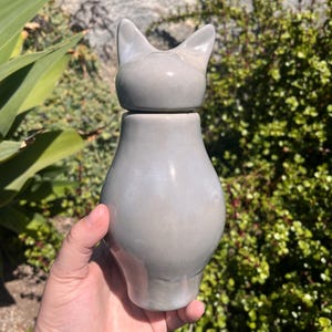 May include: A gray ceramic cat-shaped container with a lid. The container has a rounded body and a lid shaped like a cat's head with pointed ears. The container is held in a person's hand against a backdrop of green foliage.