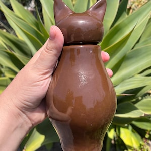May include: A brown ceramic cat-shaped container with a lid. The container has a glossy finish and a rounded body. The lid is shaped like a cat's head with pointed ears. The container is held in a hand against a background of green foliage.
