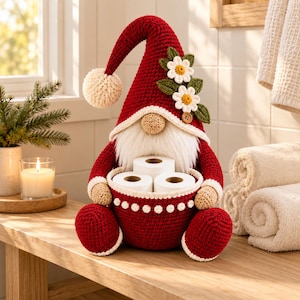 Può includere: Una statuetta di gnomo all'uncinetto in rosso scuro con un cappello alto a punta, un pompon bianco e due fiori di margherita bianchi con foglie verdi. Lo gnomo ha un naso beige, una barba bianca e tiene un cestino all'uncinetto rosso contenente tre rotoli di carta igienica.