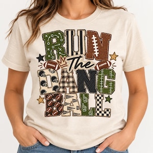 Op de afbeelding: Een crèmekleurig t-shirt met een grote afbeelding met de tekst "RUN THE DANG BALL". De letters zijn gevuld met diverse patronen, waaronder football-texturen, ruiten, strepen en sportmotieven in de kleuren groen, bruin en zwart.