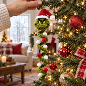Op de afbeelding: Een gehaakt groen figuurornament met een fronsende uitdrukking, voorzien van een rood-witte kerstmuts. Het ornament heeft een lang, spiraalvormig lichaam dat is versierd met afwisselend kleine rode en witte gehaakte bolletjes die verticaal hangen.