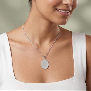 Può includere: Una collana con ciondolo ovale color argento caratterizzata da una piastra centrale liscia e tinta unita, incorniciata da un bordo metallico lucido. Il ciondolo pende da una sottile catenina color argento abbinata che poggia sulla clavicola.