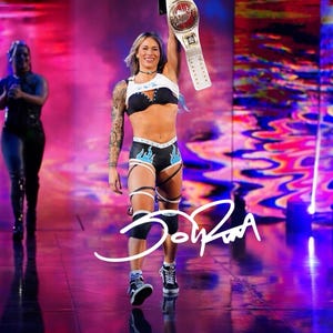 May include: A professional wrestler with blonde hair and arm tattoos wearing a white crop top, black sports bra, and black athletic shorts with blue flame accents, holding a gold and white championship belt high above their head.