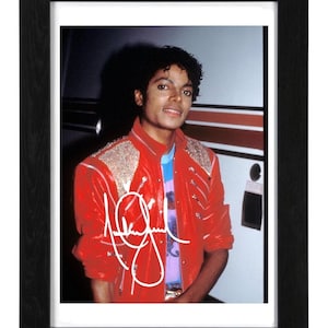 Op de afbeelding: Een ingelijste foto met een portret van Michael Jackson. Hij draagt een rode jas met zilveren versieringen en een kleurrijk shirt. De foto is wit gesigneerd en in een zwarte lijst.