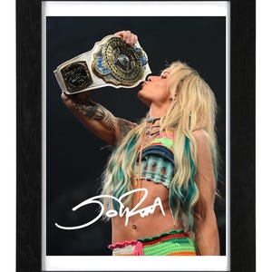 May include: A framed photograph featuring a blonde individual with teal-streaked hair kissing a metallic championship belt. The individual wears a colorful crochet top and skirt. A white signature is overlaid on the lower portion of the image.