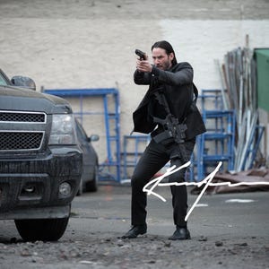 May include: A black Chevrolet Tahoe SUV with a New York license plate DQF-8093 parked next to a person in a black suit holding a black handgun and a rifle. The person is in a crouched stance, aiming the handgun forward in an outdoor setting.