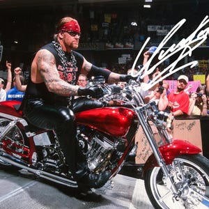 May include: A red motorcycle with chrome detailing. The rider wears a black sleeveless top, black leather trousers, and a red bandana. The motorcycle is in front of a crowd of people.