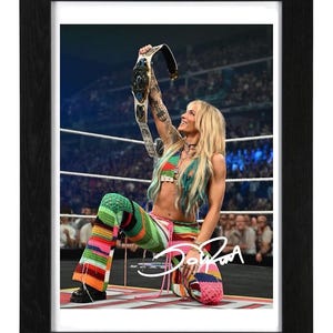 May include: A framed photograph of a blonde wrestler kneeling in a ring, holding a championship belt overhead. The wrestler wears a colorful, multi-striped crochet top and matching flared pants. A white signature is visible on the lower right.