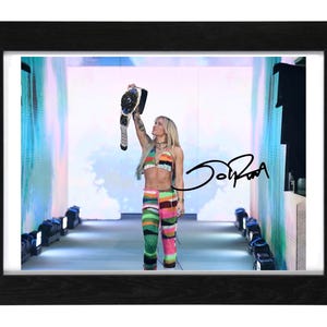 May include: A framed photograph featuring a person with blonde hair wearing a multi-colored crochet top and matching pants, holding a championship belt high in the air. The image includes a black signature overlay on the right side of the frame.