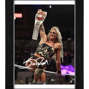 May include: A framed photograph of a professional wrestler with blonde hair, wearing a camouflage-patterned top and shorts, holding a championship belt high in the air while sitting on a ring rope. A white autograph is visible across the lower section.