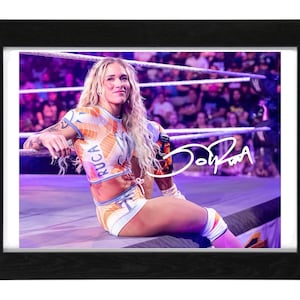 May include: A framed photograph of a wrestler with long blonde hair, wearing an orange and white patterned two-piece outfit with the word RUCA printed on the top. The image features a white signature overlay and is set in a black wooden frame.