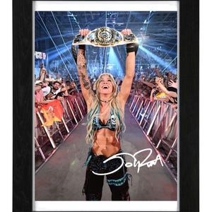 May include: A framed photograph of a wrestler with long blonde hair and tattoos on their arms, holding a championship belt high above their head in an arena. The wrestler wears a black top with a skull graphic and black pants. A white signature is visible.