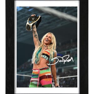 May include: A framed photograph of a blonde wrestler with long hair, wearing a colorful crocheted top and pants, holding a championship belt high in the air. A white signature is overlaid on the image, which is encased in a black wooden frame.
