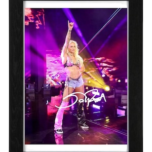 May include: A framed photograph of a person standing on a stage with bright purple lighting, wearing a dark top, light blue denim shorts, and mismatched leg coverings in pink and black, with a white signature overlaid on the image.