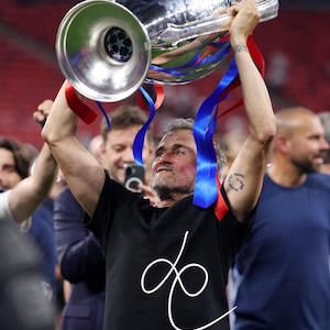 Pode incluir: Um troféu prateado com fitas vermelhas e azuis é erguido por uma pessoa que usa uma camiseta preta com um desenho branco. O troféu tem uma tigela grande e uma base cilíndrica. Outras pessoas são visíveis.