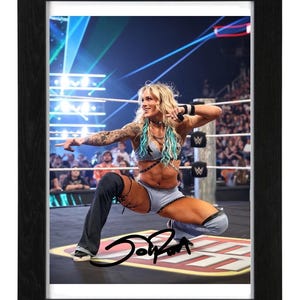 May include: A framed photograph of a professional wrestler in a ring, wearing a light blue two-piece outfit with black straps and one long pant leg. The wrestler has blonde hair with teal streaks, tattoos on the arm, and a black signature across the bottom.