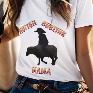 May include: White t-shirt with the text "Mutton Busting Mama" in a Western-style font. The shirt features a graphic of a child riding a sheep. A woman wearing a cowboy hat smiles next to a horse.