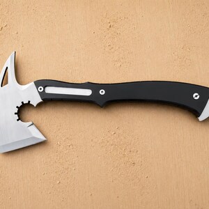 May include: A tactical hatchet featuring a silver-colored metal blade with a pointed top spike, a circular cutout, and a curved black handle. The handle includes a silver inlay, two visible screws, and a lanyard hole at the end of the grip.
