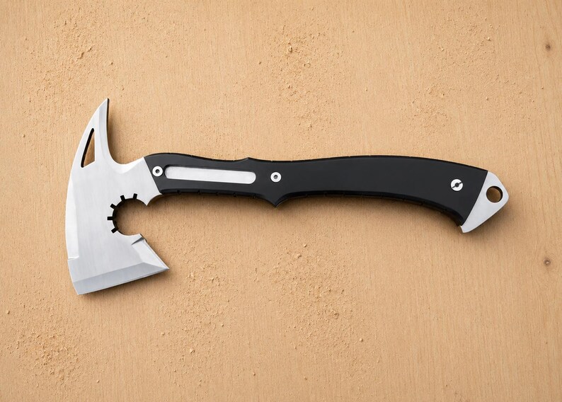 May include: A tactical hatchet featuring a silver-colored metal blade with a pointed top spike, a circular cutout, and a curved black handle. The handle includes a silver inlay, two visible screws, and a lanyard hole at the end of the grip.