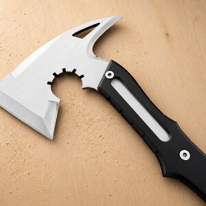 May include: A tactical hatchet featuring a silver-colored metal head with a sharp blade, a curved cutout, and a pointed spike. The handle is black with a central silver inlay and is secured by two visible silver screws near the axe head.
