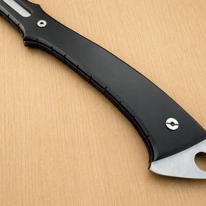May include: A curved knife handle featuring a smooth matte black finish with a series of small vertical notches along the edge. The handle is secured with two silver-toned circular screws and terminates in a metallic pommel with a circular hole.
