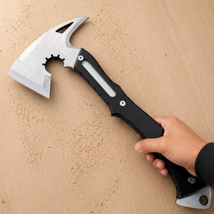 May include: A tactical hatchet featuring a silver-colored metal head with a curved blade, a cutout design, and a notched circular opening. The handle is black with a silver-colored inlay, secured by two visible screws, and includes a lanyard hole.