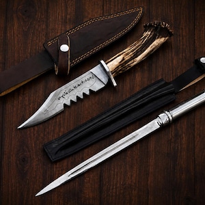 May include: A hunting knife with a serrated blade featuring etched script and an antler handle, paired with a brown leather sheath. Beside it lies a slender, double-edged metal dagger with a polished silver handle and a black leather sheath.