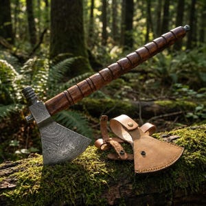 Könnte beinhalten: Ein handgefertigtes Tomahawk mit einer gemusterten Stahlklinge und einem dunklen Holzgriff. Der Griff weist geschnitzte Details auf. Eine hellbraune Lederscheide schützt die Klinge. Das Tomahawk liegt auf einem moosbewachsenen Baumstamm im Wald.