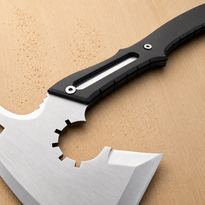 May include: A tactical axe featuring a brushed stainless steel head with a circular cutout containing gear-like teeth. The handle is matte black with a silver inlay and is secured by two visible hex screws, showing a modern, industrial design.