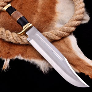 May include: A large, polished steel hunting knife with a curved blade. The handle features a black and brown grip with gold-colored accents. The knife is displayed on a brown and white fur surface.