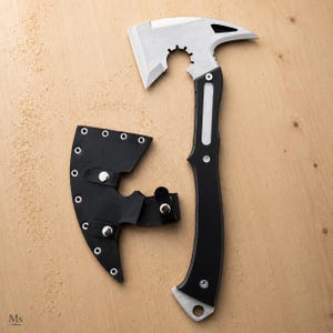 May include: A tactical hatchet with a silver-toned metal head featuring a notched blade and a pointed spike, attached to a black handle. A matching black leather sheath with silver metal rivets and snap closures is positioned next to the hatchet.