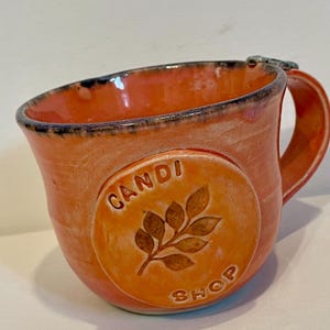 May include: Orange ceramic mug with a handle and a decorative circular emblem. The emblem features the text "Candi Shop" and a leaf design. The mug has a dark blue rim and a textured surface.