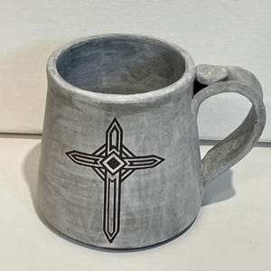 May include: Gray ceramic mug with a cross design. The mug has a wide, cylindrical shape with a handle and a decorative cross graphic on the front. The mug is a light gray color.
