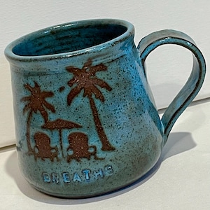 May include: A ceramic mug with a turquoise glaze and a brown palm tree and beach scene. The mug features two palm trees, two beach chairs, and the word "BREATHE" in a lower case font.